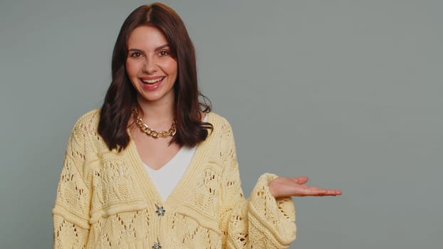 Lovely woman in cardigan showing thumbs up and pointing empty place, advertising area for commercial text, copy space for goods promotion. Young pretty girl. Indoor studio shot on gray wall background