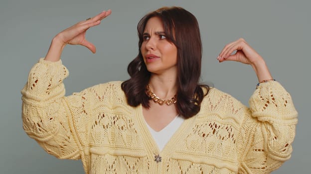 Bored pretty young woman in cardigan showing blah blah gesture with hands, has not interested in the conversation, gossips, empty promises, lier. Millennial girl isolated on gray studio background