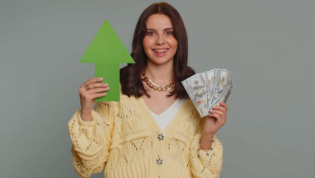 Happy pretty woman winner holding arrow sign pointing up, career growth and money dollar exchange increase. Young adult girl showing up indicating successful development progress on gray background
