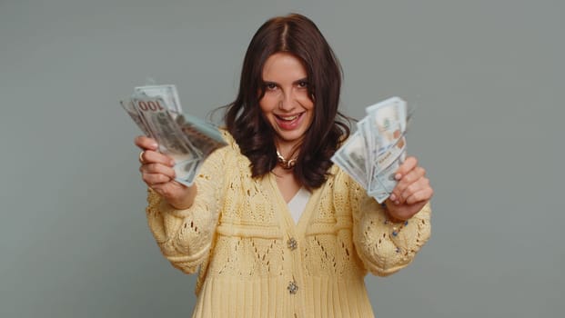 Rich pleased boss millennial woman waving money dollar cash banknotes bills like a fan, success business career, lottery winner, big income, wealth. Young pretty girl isolated on gray background