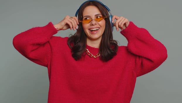 Happy pretty woman in headphones listening music and dancing disco fooling around having fun expressive gesticulating hands, relaxing on party. Caucasian girl isolated on gray studio wall background