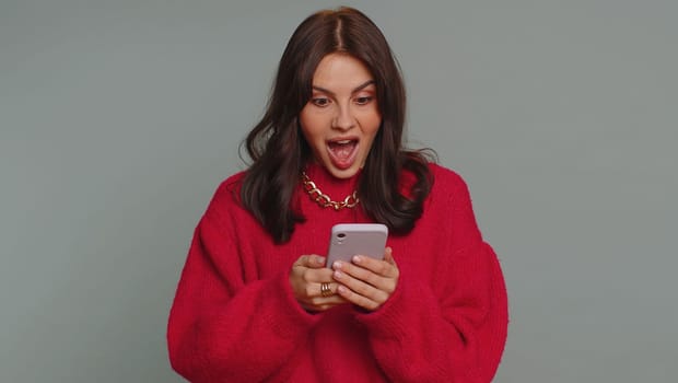 Happy excited joyful pretty woman in sweater use mobile cell phone typing browsing shouting say wow yes found out great big win, good news, lottery goal achievemen, celebrating success, winning