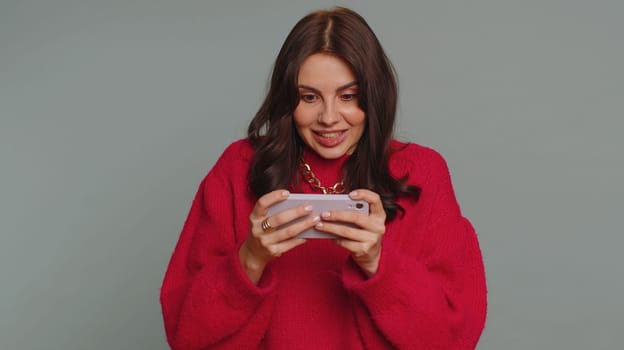 Worried funny addicted woman enthusiastically playing racing video games on mobile phone. Young pretty girl using smartphone gadget app with drive simulator isolated on gray studio background