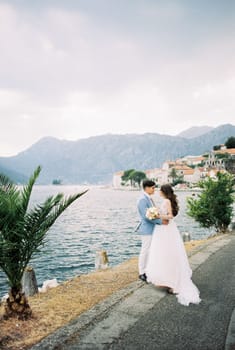 Bride and groom stand hugging on the embankment of Perast against the backdrop of the mountains. Montenegro. High quality photo
