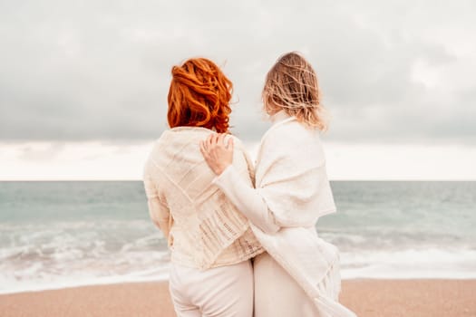 Women sea walk friendship spring. Two girlfriends, redhead and blonde, middle-aged walk along the sandy beach of the sea, dressed in white clothes. Against the backdrop of a cloudy sky and the winter sea. Weekend concept