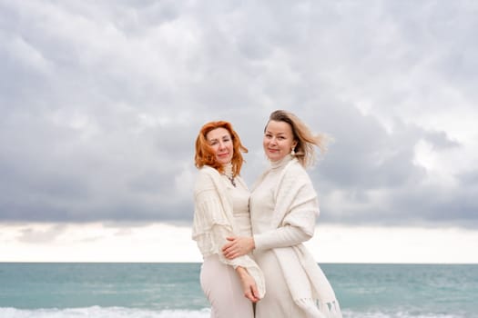 Women sea walk friendship spring. Two girlfriends, redhead and blonde, middle-aged walk along the sandy beach of the sea, dressed in white clothes. Against the backdrop of a cloudy sky and the winter sea. Weekend concept