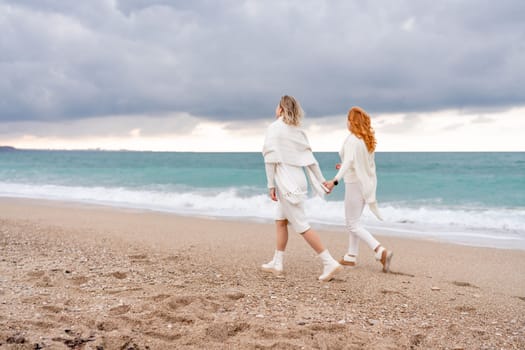 Women sea walk friendship spring. Two girlfriends, redhead and blonde, middle-aged walk along the sandy beach of the sea, dressed in white clothes. Against the backdrop of a cloudy sky and the winter sea. Weekend concept