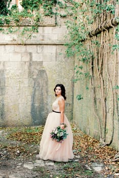 Bride with a bouquet stands in the courtyard of an old stone building, entwined with ivy. High quality photo