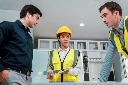 A team of competent engineers wearing safety equipment is working on blueprints with a tablet while also discussing with the investor in the office.