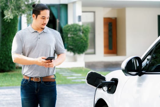 Progressive asian man install cable plug to his electric car with home charging station in the backyard. Concept use of electric vehicles in a progressive lifestyle contributes to clean environment.