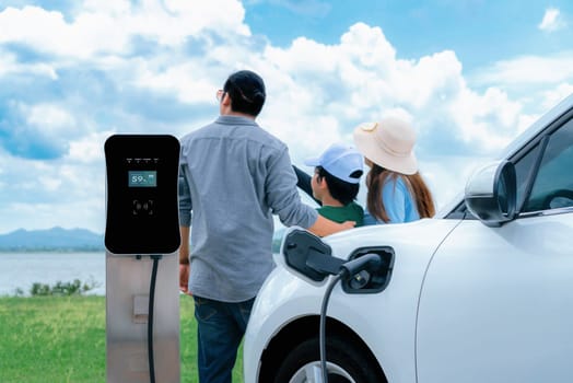 Concept of progressive happy family with electric vehicle enjoy their time at natural outdoor scenic with lake, greenfield and cloudscape background. Electric vehicle driven by green renewable energy.