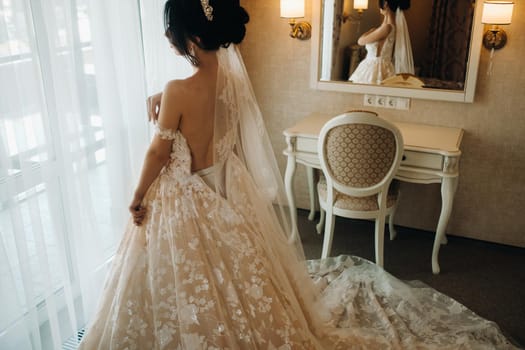 The bride in a beautiful dress with a long train stands near the window in the interior..