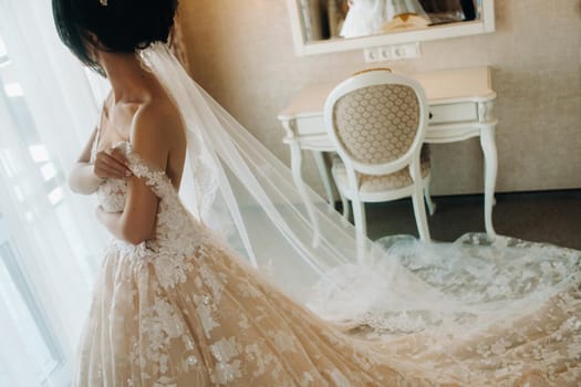 The bride in a beautiful dress with a long train stands near the window in the interior..