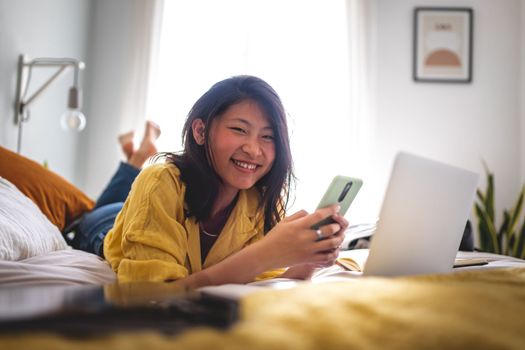 Teenage asian girl college student using mobile phone lying on bed while doing homework using laptop looking at camera. Lifestyle, technology and education concept.