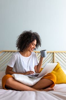 Working at home. African American young woman sitting on bed drinking hot coffee while typing on laptop. Relaxing in bedroom. Vertical image Home concept.