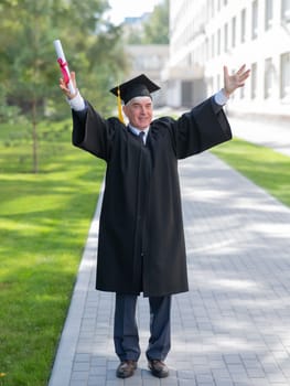 An elderly gray-haired man in a graduate robe spread his arms to the sides and holds a diploma outdoors. Vertical