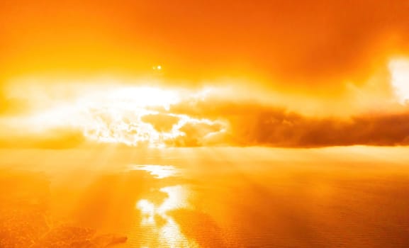 A red burning sunset with the silhouette of a cliff over the sea. Abstract nature summer or spring ocean sea background. Small waves on golden warm water surface with bokeh lights from sun.