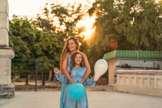 Mother daughter family sunset. Portrait of mother and daughter in blue dresses with flowing long hair against the backdrop of sunset. A woman hugs and presses the girl to her, holding balloons in her hands.