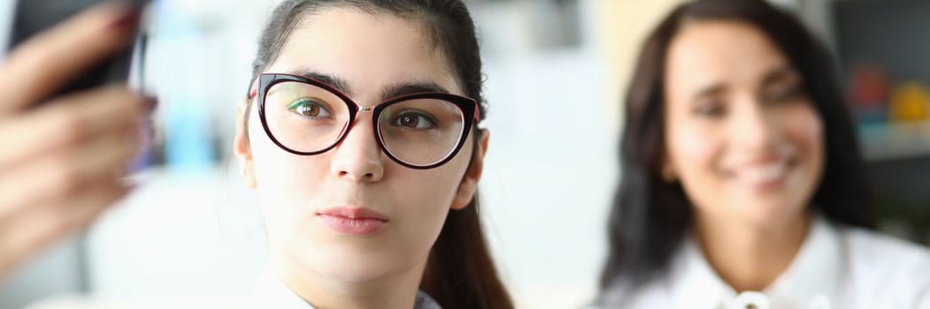 Young woman in glasses holds phone in front of face and smiles at colleagues in office. Video call or selfie at work