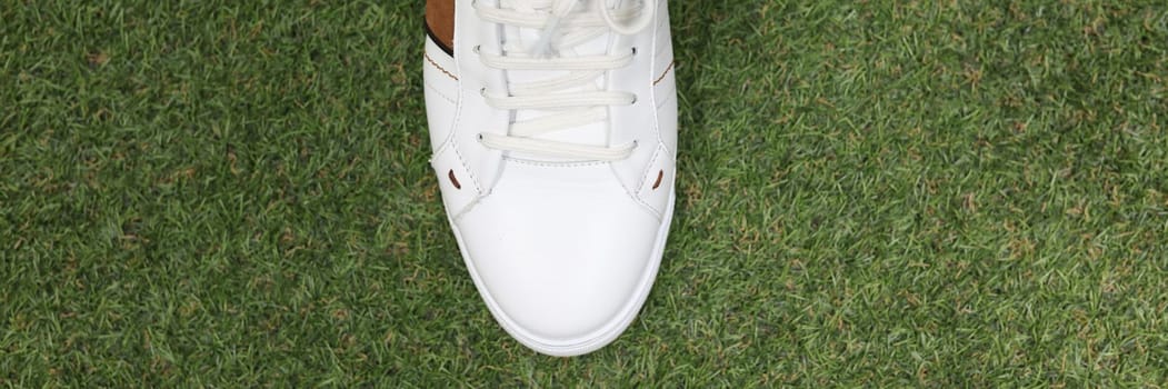 Man is wearing white sneakers on green lawn grass. Stylish trendy sports sneakers