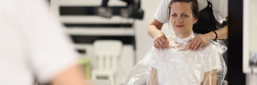 Female hairdresser puts paper collar bag around woman neck to protect clothes she cuts hair indoors. Caucasian beautiful lady changes image in a hairdresser