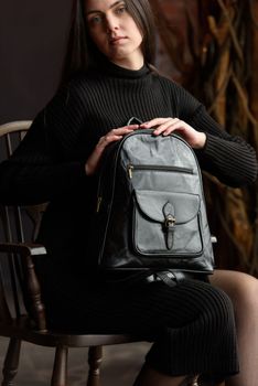 a brunette girl in a knitted black dress poses while sitting with a shiny black leather backpack in her hands