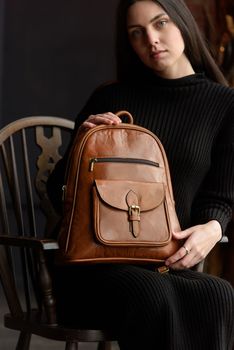 a brunette girl in a knitted black dress poses while sitting with a shiny brown leather backpack in her hands