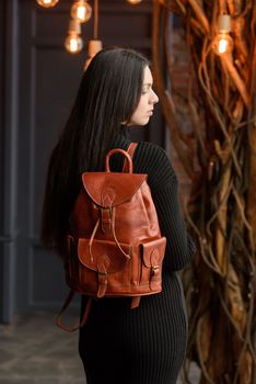 a brunette girl in a knitted black dress poses while standing with a shiny red leather backpack in her hands