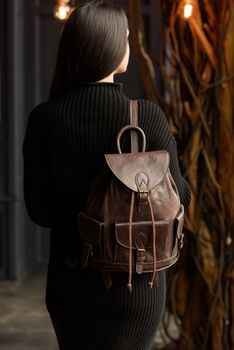 a brunette girl in a knitted black dress poses while standing with a shiny brown leather backpack in her hands