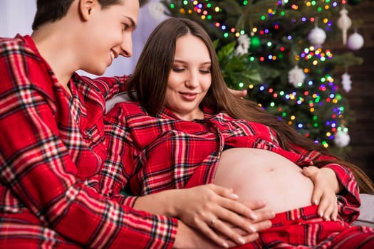 A couple expecting a baby sits on a couch near a dressed Christmas tree, which can be seen in a blurry background. The parents-to-be are wearing the same plaid pajamas.