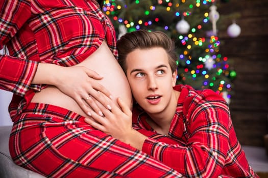 Pregnant belly seen in close-up in profile. A man puts his ear to the pregnant belly. The man has his mouth open and listens while holding his hand on his partner's belly.