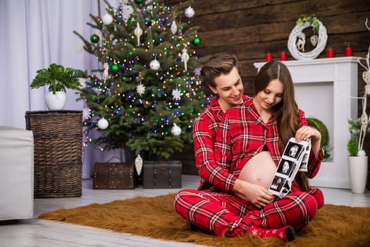 A couple sitting on a fluffy carpet is looking at usg photos showing their baby. The woman is advanced in pregnancy and exposes her naked pregnant belly. The man is sitting behind her back.