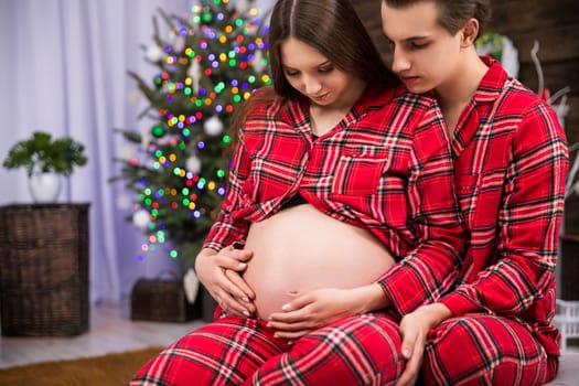 A couple expecting a baby sits hugging. The woman leans against the man with her back and exposes her pregnant belly. The man hugs the woman and strokes her pregnant belly.