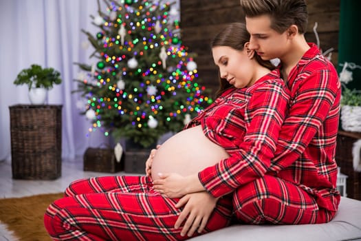 A pregnant woman leans with her back against a man sitting behind her. The man hugs the woman and holds his hand on her stomach. The hugging couple can be seen in profile against a smudged Christmas tree.