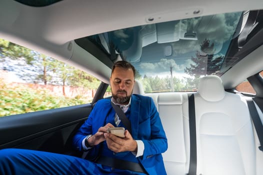 A caucasian man in a blue suit uses a smartphone in the back seat of a car. Business class passenger