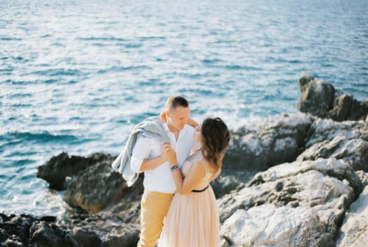 Bride hugs groom by the neck on the rocks by the sea. High quality photo
