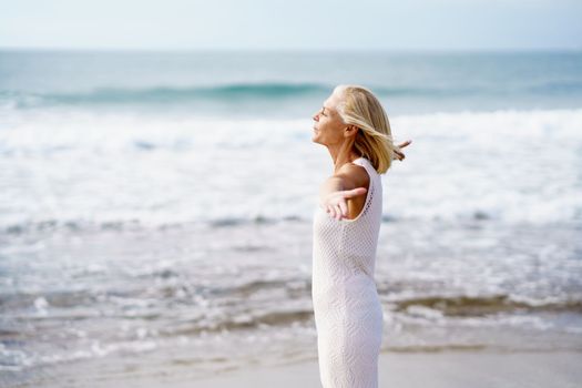 Mature woman opening her arms on a tropical beach, spending her leisure time. Elderly female enjoying her retirement at a seaside retreat.