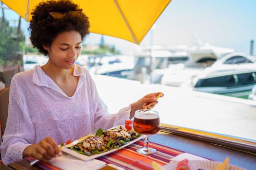 Glad young black woman in white blouse with curly hair smiling and eating tasty exquisite dish, while sitting at table on blurred background of embankment on sunny summer day on resort.
