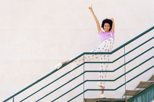 Full body of happy African American female, in casual outfit pointing up with raised arm and looking at camera while standing on stairway with leg on railing against white background