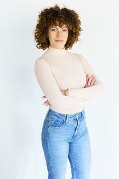 Self assured female model with dark curly hair in beige turtleneck and jeans crossing hands while standing against white background