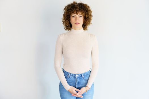 Portrait of emotionless adult curly haired woman in turtleneck and jeans standing with back straight and hands clasped looking at camera against white background