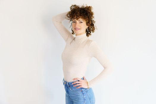 Adult curly haired female in casual wear standing with hand on hip and touching hair while looking at camera against white background