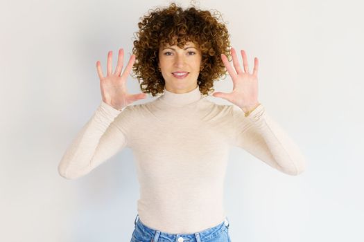 Attractive curly haired female in casual outfit raising hands and showing number ten while looking at camera on white background