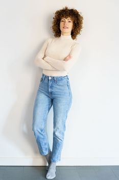 Full body of curly haired female in jeans and beige turtleneck leaning on white wall with hands crossed looking at camera