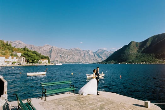 Bride hugs groom from behind on the pier near the bench. High quality photo