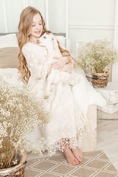 A blonde girl with long hair is sitting on the couch with a goat at home. The concept of animal care