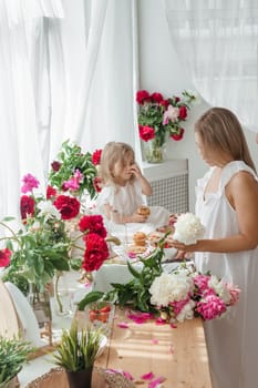 A little blonde girl with her mom on a kitchen countertop decorated with peonies. The concept of the relationship between mother and daughter. Spring atmosphere.