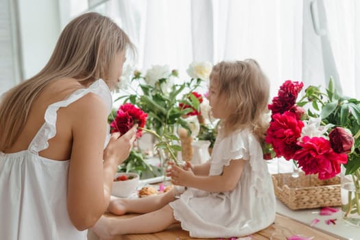 A little blonde girl with her mom on a kitchen countertop decorated with peonies. The concept of the relationship between mother and daughter. Spring atmosphere.