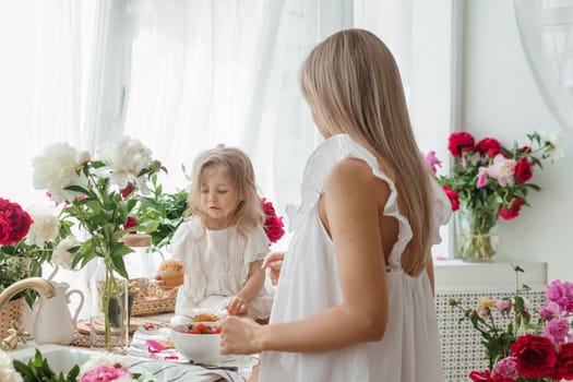 A little blonde girl with her mom on a kitchen countertop decorated with peonies. The concept of the relationship between mother and daughter. Spring atmosphere.