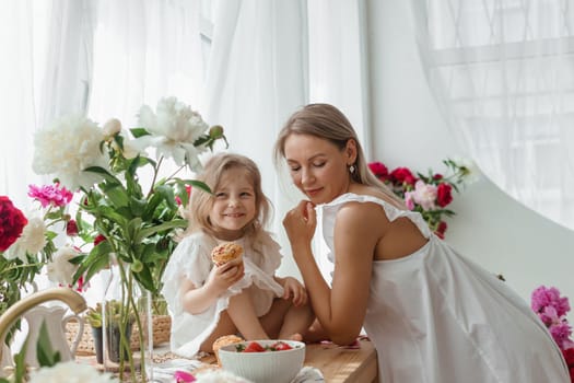 A little blonde girl with her mom on a kitchen countertop decorated with peonies. The concept of the relationship between mother and daughter. Spring atmosphere.